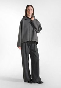 Felpa grigia oversize con zip frontale e ampio cappuccio, abbinata a pantaloni larghi grigi con motivo e scarpe nere.