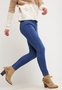 Vaqueros pitillo azules de talle alto combinados con botines beige con detalles de flecos y tacón de bloque. El suéter es de punto multicolor.