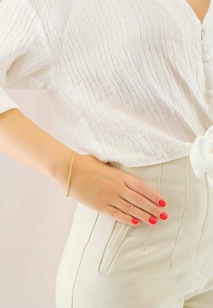 Goldene gedrehte Armbanduhr, die am Handgelenk getragen wird, vor einer weißen strukturierten Bluse und cremefarbenen Hosen. Helle rote Nägel mit Nagellack.