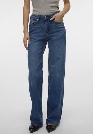 Femme portant un jean bleu taille haute à jambes larges, un haut gris sans manches et des chaussures noires pointues avec détail boucle, debout avec une main dans la poche.