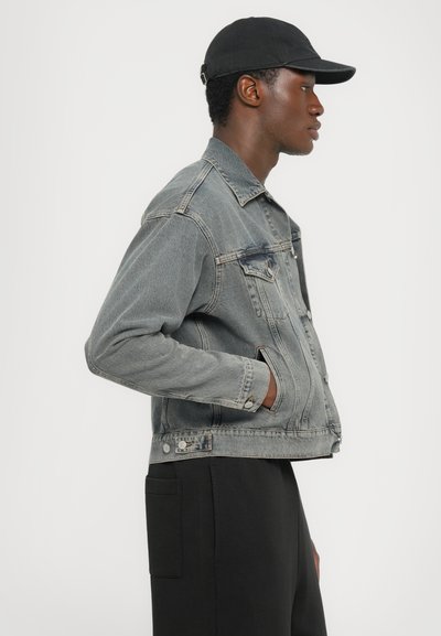 Jachetă din denim de culoare gri-albastră estompată, cu un croi clasic, închidere cu butoane, buzunare la piept și un finisaj texturat. Asortată cu pantaloni negri.