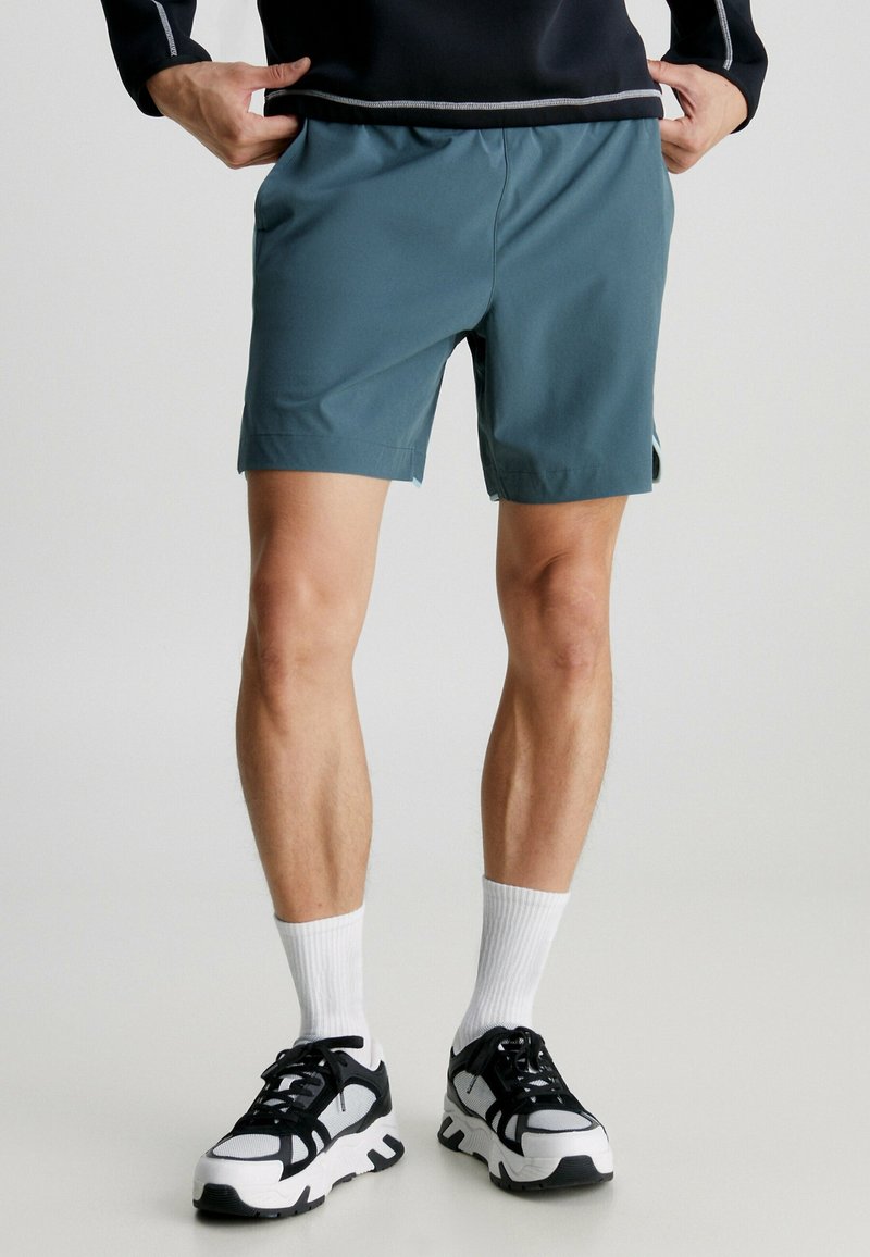 Personne portant un short de sport bleu sarcelle, des chaussettes blanches, des baskets noires et blanches et un haut noir, les mains dans les poches, debout à l'intérieur.