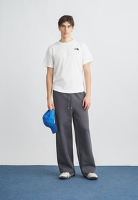 Λευκό βαμβακερό t-shirt με μαύρο λογότυπο, γκρι φαρδιά φόρμα, μπλε καπέλο μπέιζμπολ κρατημένο στο χέρι και γκρι αθλητικά παπούτσια. Απλό φόντο.