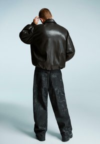 Giacca bomber in pelle nera con finitura lucida, forma oversize e texture morbida. Abbinata a pantaloni scuri, larghi e leggermente strutturati.