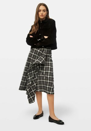 Veste en velours côtelé noir, jupe midi à carreaux noirs et blancs avec un détail drapé, et chaussures plates noires avec un accent en noeud.