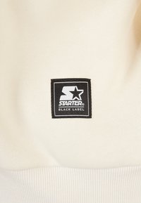 Kremasti dukserica s crnim kvadratnim aplikacijom i bijelim natpisom "STARTER BLACK LABEL" te rebrastim manžetama.