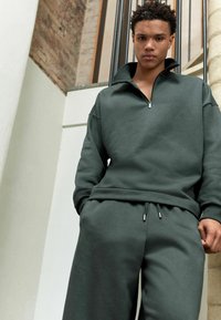 Sudadera de cremallera en verde oscuro con cuello alto y corte relajado, combinada con pantalones de chándal a juego que cuentan con cordón y detalles en metal plateado.
