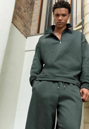Dark green zip-up sweatshirt with high collar and relaxed fit, paired with matching sweatpants featuring drawstring and silver hardware accents.