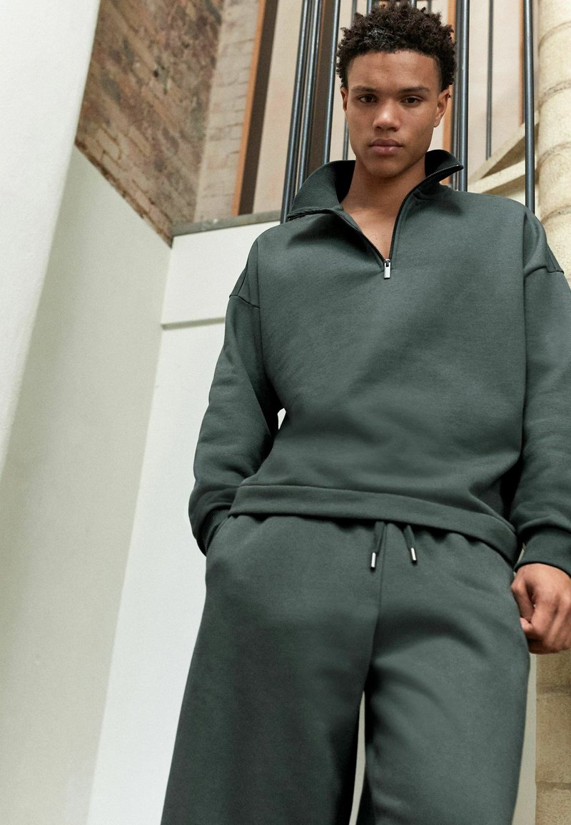 Sudadera de cremallera en verde oscuro con cuello alto y corte relajado, combinada con pantalones de chándal a juego que cuentan con cordón y detalles en metal plateado.