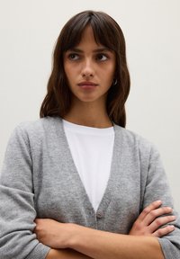 Cardigan grigio con design a V, texture morbida e chiusura con un solo bottone, indossato sopra una maglietta bianca. Braccia incrociate in una posa neutra.