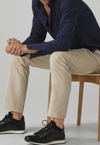 Man in een marineblauw shirt, beige broek en zwarte sneakers zit op een houten stoel met zijn handen gevouwen, tegen een lichtgrijze achtergrond.