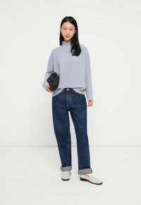 Svijetlosivi pulover s visokim ovratnikom, uparen s tamnoplavim, širokim farmerkama s podvrnutim rubovima, i svijetlim slip-on cipelama. Drži crnu clutch torbicu.
