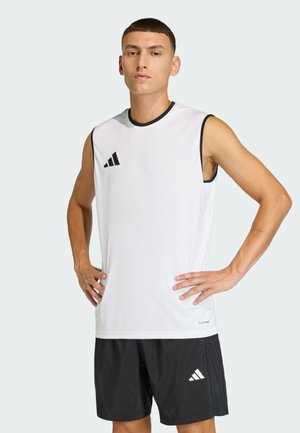 Jeune homme debout, mains sur les hanches, portant un t-shirt de sport blanc sans manches et un short noir avec logos Adidas sur fond clair.