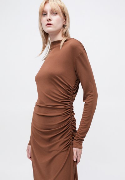 Brun langermet kjole med rynkede detaljer langs sidene. Laget av mykt, stretchy stoff. Rund hals og kroppsnær silhuett.