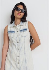 Denimklänning i ljusblått, utan ärmar med knappfront, bröstfickor och en blekt tvätt. Modellen bär svarta solglasögon.