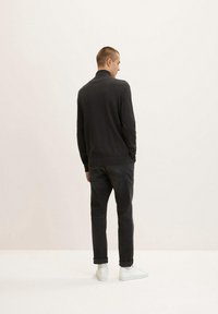 Černý textured pulover s vysokým límcem, nošený s tmavými džíny a bílými teniskami. Pohled zezadu zdůrazňuje střih a materiál oděvu.