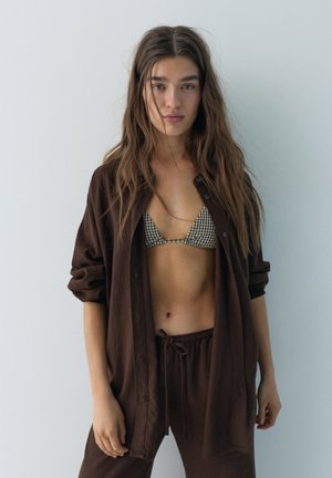 Žena s dugom smeđom kosom nosi kockasti gornji dio bikinija i labavu smeđu košulju na kopčanje s odgovarajućim hlačama s vezicama, stojeći uz bijeli zid.