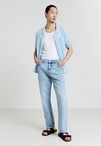 Camicia azzurra a maniche corte, top bianco senza maniche, jeans in denim azzurra a gamba dritta, sandali neri con suola texturizzata, postura eretta.