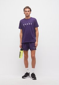 Νεαρός άνδρας που φοράει μοβ αθλητικά σορτς και μπλούζα με την επωνυμία HYROX, κρατάει κίτρινο παγούρι νερού PUMA, στέκεται μπροστά σε λευκό φόντο.