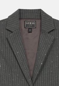 Γκρι σακάκι με λεπτές ρίγες και διακριτική λαμπερή λεπτομέρεια, γιακάς με εγκοπή και εσωτερική επένδυση. Η ετικέτα γράφει "GUESS, EST. 1981." Παρουσιάζεται ταμπέλα μεγέθους 8.