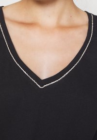 Černý top s výstřihem do V z jemného materiálu, zdobený stříbrnými flitry podél výstřihu, který zvýrazňuje hladkou texturu a moderní design.