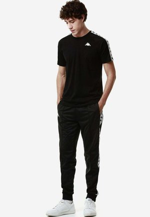 Jeune homme regardant vers le bas, portant un t-shirt noir Kappa et un pantalon de survêtement assorti avec des baskets blanches, debout les mains dans les poches.