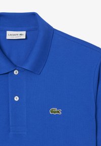 Kirkas sininen polo-paita, jossa on teksturoitu kangas, kaksi nappia ja pieni vihreä krokotiililogo vasemmalla puolella lähellä kaulusta.