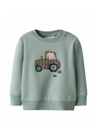 Hellgrüner Sweatshirt mit gerippten Bündchen und Saum. Mit einer Kuh-Illustration in einem braunen Traktor, darunter schwarze Reifenabdrücke.