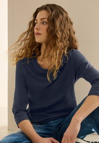 Mörkblå, långärmad topp med rund halsringning och mjuk textur, tillsammans med ljusblå denimjeans. Spröda lockar ramar ansiktet.