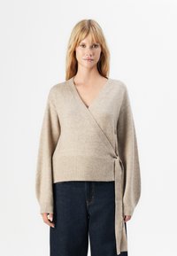 Blond kvinna i en beigefärgad omslagströja knuten i sidan och mörkblå högmidjade jeans, står mot en vit bakgrund.