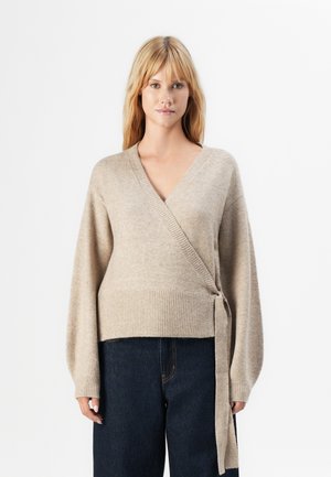 Mujer rubia vestida con un suéter beige de envoltura atado de lado y jeans de talle alto en azul oscuro, de pie frente a un fondo blanco.
