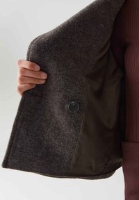 Grijze wolmix jas met een bruine satijnen voering. Voorzien van een knoopsluiting, gestructureerde schouders en een gladde, textuurgegeven buitenkant.