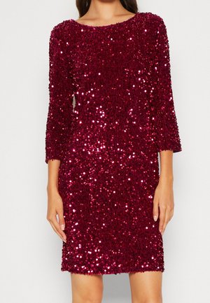 Femme portant une robe à manches longues, longueur genou, rouge foncé à sequins avec un col rond, debout les bras relâchés le long du corps.
