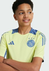 Maillot de football jaune à manches courtes avec des accents bleus, portant le logo de l'Ajax et trois étoiles au-dessus. Confectionné en tissu léger.