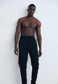 Zwarte joggings met rode zijstrepen, trekkoord in de taille en een zachte textuur. Model poseert zonder shirt tegen een neutrale achtergrond.