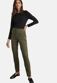 Zwarte lange mouwen top gecombineerd met hoogwaist olijfgroene broeken met voornaad en zijzakken. Model draagt zwarte instappers.