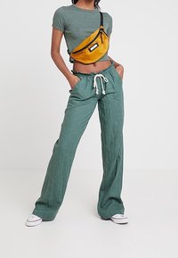 Pantalon long vert, ample, avec tissu texturé et taille à cordon de serrage. Porté avec un crop top côtelé gris et une sacoche jaune à la taille.