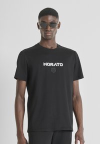 Camiseta negra de algodón, presenta el logo "MORATO" en blanco con un emblema texturizado en negro debajo, cuello redondo, mangas cortas, corte relajado.