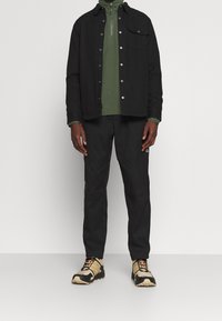 Schwarze Button-Up-Jacke über einem grünen Zip-Kragen-Oberteil, kombiniert mit schwarzen Hosen und tanfarbigen und schwarzen Sneakers mit orangen Akzenten.