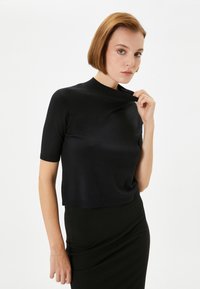 Sort kortærmet bluse med høj hals og ribbet kant, lavet af et glat, let materiale. Båret sammen med en tætsiddende sort nederdel.