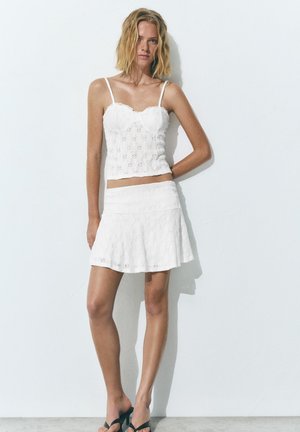 Frau in weißem ärmellosem Top aus Lochspitze und passendem ausgestelltem Rock, stehend an einer weißen Wand, trägt schwarze Sandaletten mit Absatz.