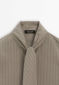 Chemise beige à fines rayures avec un col large et un détail de nœud lâche au niveau du cou, étiquetée Massimo Dutti, fabriquée au Maroc.