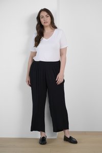 Weißes kurzärmliges V-Ausschnitt-Shirt, kombiniert mit locker sitzenden schwarzen Hosen. Die Hose hat eine entspannte Silhouette und einen elastischen Bund. Dazu werden schwarze Slipper getragen.