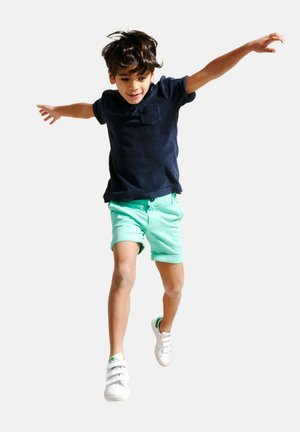 Marineblauwe poloshirt, mintgroene shorts, witte sneakers. Kind springt met uitgestrekte armen, toont de levendige kleuren en de relaxte pasvorm van de outfit.