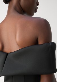 Robe noire à épaules dénudées en tissu satin lisse, avec un détail de nœud structuré et une silhouette ajustée avec une texture subtile.