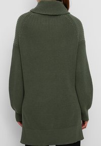 Femme portant un long pull épais en tricot vert olive avec épaules tombantes et poignets côtelés, debout de dos sur un fond uni.