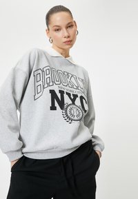 Sudadera gris con un diseño impreso en negro que presenta "BROOKLYN" y "NYC". Puños y cuello acanalados, corte relajado, textura suave.
