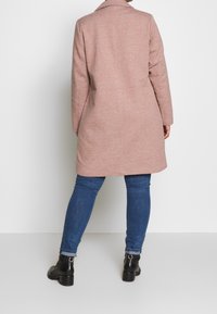 Pitkä vaaleanpunainen villatakki, jossa on lovellinen kaulus, reilu malli ja teksturoitu kangas, yhdistettynä sinisiin farkkuihin ja mustiin nilkkureihin.