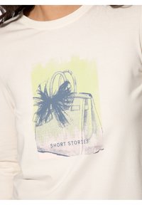 Crème korteboven t-shirt met een bedrukt handtasontwerp met een zwarte strik en de tekst "SHORT STORIES" in het blauw op een lichtgroene achtergrond.