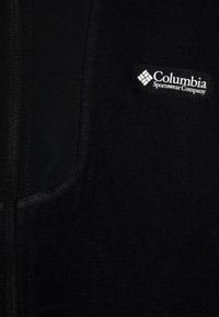 Gilet en polaire noire avec fermeture éclair à l'avant, comportant une poche en tissu et un logo blanc de la Columbia Sportswear Company. Texture douce, design simple.